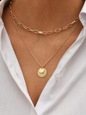 Colliers en chaîne dorée avec pendentif rond texturé et collier chaîne en maillons ovales, élégants et modernes, portés ensemble sur une peau caramel avec chemise blanche ouverte, parfaits pour un look chic et tendance.